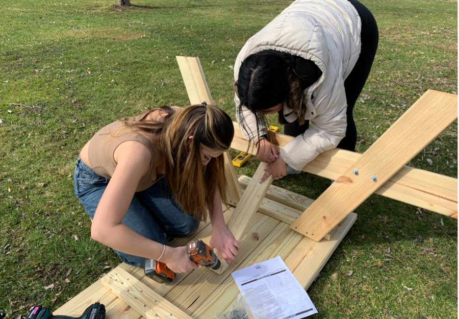 two young teens work outside in winter coats using a drill to put together a picnic table